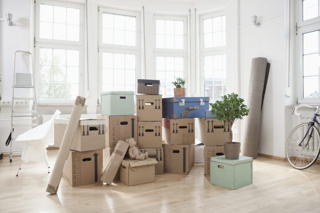 Stacked moving boxes and packed household items arranged in a bright, empty room with large windows, including a plant, rolled rug, and packing supplies ready for relocation.