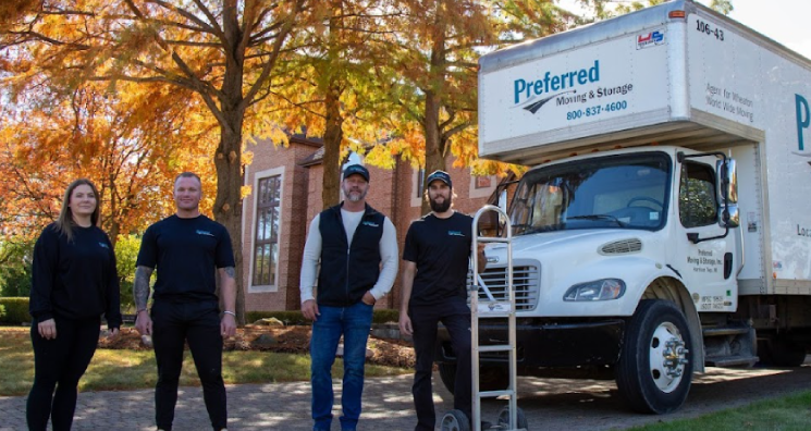 Team of Preferred Moving & Storage movers standing in front of a company truck outside a residential home with autumn trees in the background.