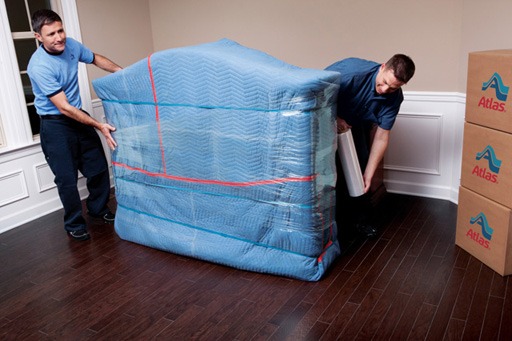 Professional movers wrapping and lifting a large piece of furniture in blue protective padding inside a home, with stacked moving boxes nearby.

