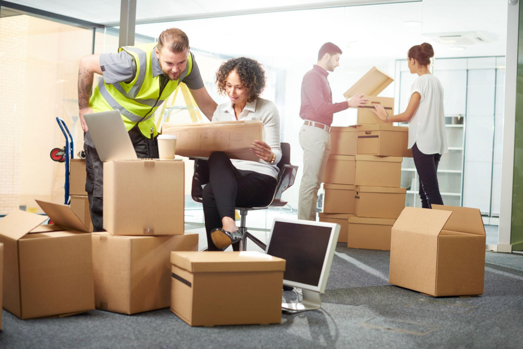 Professional office movers and employees coordinating a commercial business relocation with packed boxes and office equipment in a modern workspace.